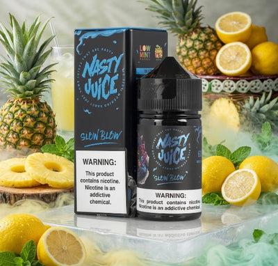 Nasty Juice Glow Blow e-liquid bottle and its retail box displayed on a reflective surface, surrounded by fresh pineapples, lemons, mint leaves, and a glass of yellow drink in a styled product scene.