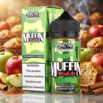 A bottle and box of One Hit Wonder Muffin Man e-liquid vape juice, styled with muffins, apples, and cinnamon sticks on a reflective surface.